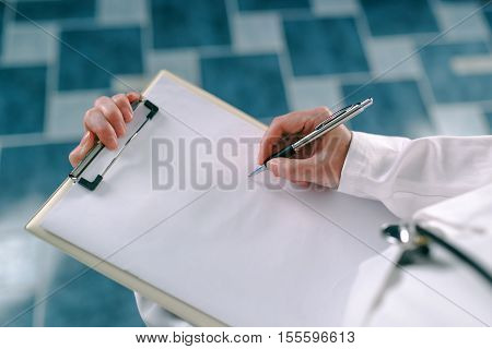 Female doctor in white uniform writing on clipboard paper patient's medical history. Woman as health specialist in exam er disease prevention visit check or healthcare lifestyle concept