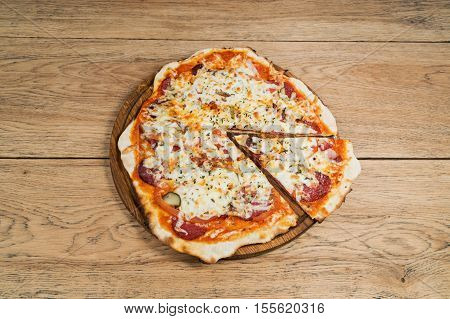 Delicious round pizza with salami and pickles with cut off piece, served on a wooden plate on old wood table