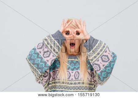 Young blonde woman in sweater looking through fingers isolated on the gray background