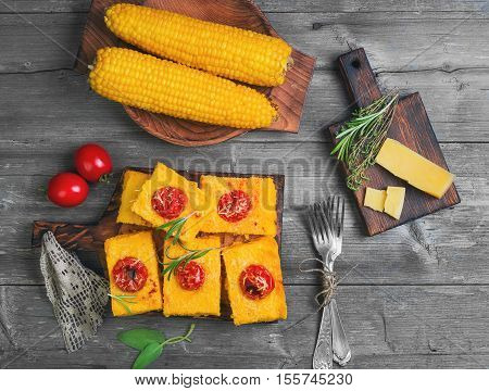 Pieces Corn polenta baked with cherry tomatoes Parmesan cheese on board. Boiled corn on wooden plate for polenta. Green for corn polenta thyme rosemary. Gray wooden background. Top view