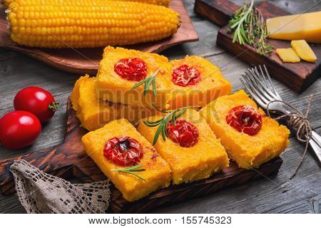 Pieces Corn polenta baked with cherry tomatoes Parmesan cheese on board. Boiled corn on wooden plate for polenta. Green for corn polenta thyme rosemary. Gray wooden background.