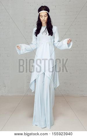 Girl child flowers, hippie in bright clothing, bottoms, shirt and armband. Model in studio posing to the photograther staying near grey bricks wall. She has beautiful white costume