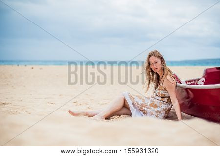 A Young Girl Sits On The Beach Leaning Against A Boat