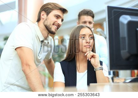 Group Of Young People In Casual Wear Sitting At The Office Desk And Discussing Something While Looki