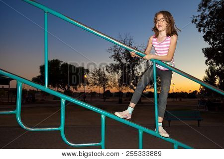 A Young Girl With Missing Teeth, Climbs Jungle Gym Bars At A Playground At Sunset.  There Is A Lot O