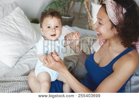 Top View Of Caring Loving Young Mixed Race Mother Hoding Her Sweet Newborn Son, Sitting On Bed And L