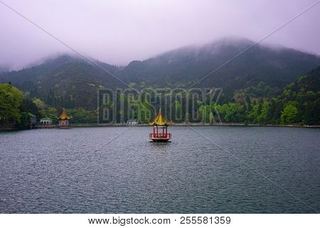 Glacial Lulin Lake View With A Chinese Pavilion In The Middle And Green Cloudy Mountains In Backgrou