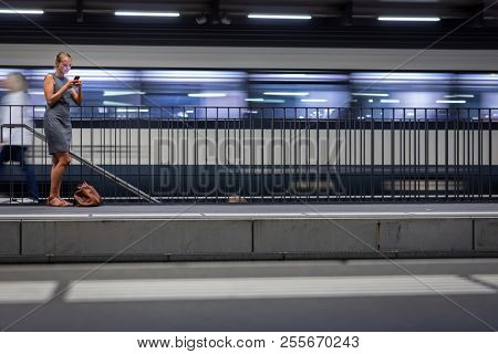 Pretty, young female commuter waiting for her daily train in a modern trainstation, using her cellphone while waiting (color toned image)