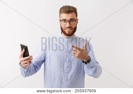 Portrait of a smiling young bearded man in eyeglasses standing isolated over white, holding mobile phone