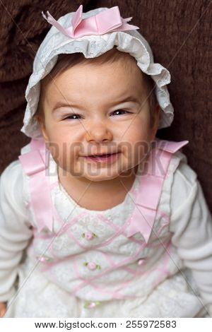 An Adorable, Baby Girl In A White And Pink Floral Outfit With A Hat Smiles With A Scrunched Face And