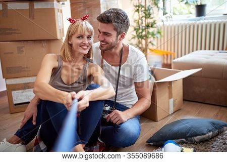young couple taking selfie  in new apartment, sitting on a floor with unpacked stuff in cardboard boxes.  new apartment, new beginning, joy, carefree, leisure  concept