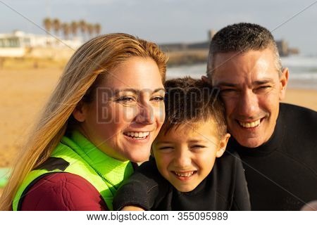 Happy Family In Wetsuits On Beach. Close-up Portrait Of Cheerful Middle Aged Parents And Cute Little