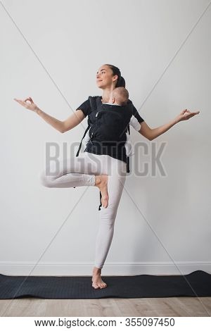 Young Mother Doing Yoga Exercises And Meditation With Her Little Baby In Ergo Backpack.
