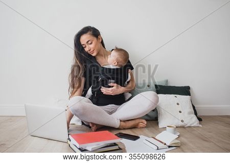 Young Freelance Mother Having Her Baby In Ergo Backpack And Working At Home Using Laptop And Smart P