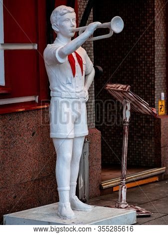The Figure Of A Pioneer With Clarion In His Hand Near Building. White Sculpture Of A Boy With A Red 