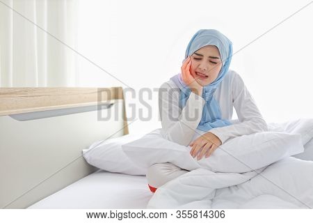 Beautiful And Young Asian Muslim Woman In Sleepwear With Tooth Pain Emotion Sitting On Bed. Cute Gir