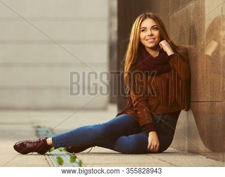 Happy young fashion woman sitting on street sidewalk  Stylish female model in brown leather fringe suede jacket and dark blue jeans