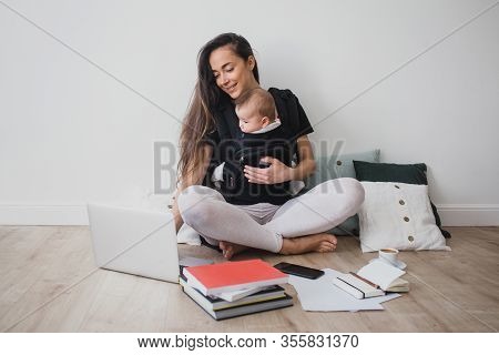 Young Freelance Mother Having Her Baby In Ergo Backpack And Working At Home Using Laptop And Smart P