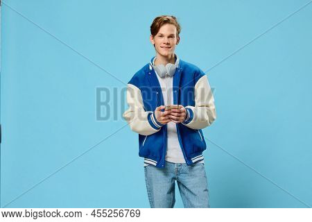 Young Man In A Blue Jacket Stands With A Phone In His Hands And White Headphones Around His Neck Wri
