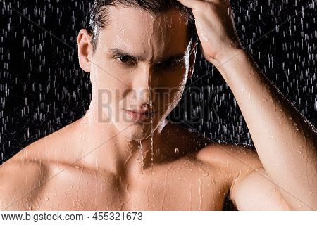 Wet Man With Naked Shoulders Touching Hair And Looking At Camera Under Shower On Black Background