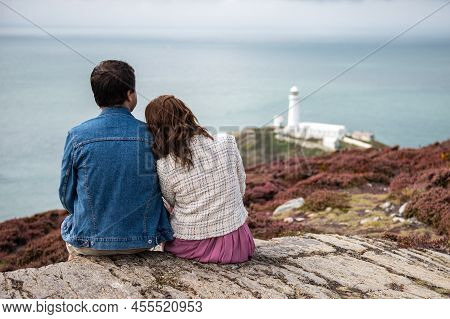 Couple Is Sitting On The Edge Of Cliff, South Stack Lighthouse On Background, Wales, Anglesey. Stayc