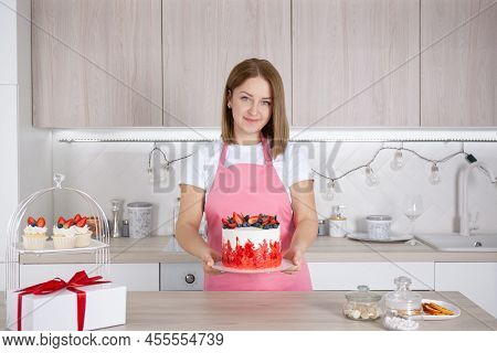 Pastry Chef Confectioner Young Caucasian Woman In The Kitchen. She Holds Out The Cake On A Plate. Ne