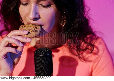 Young Female Blogger Sitting In Studio In Pink Neon Light Recording Crunchy Cookies Eating Sounds Fo