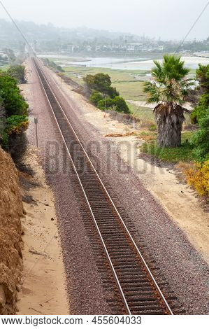 Landscape With Empty Train Tracks In Southern California With A Town On The Background And Palm Tree