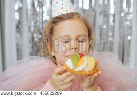 Oh, How Delicious. Pretty 4-5-year-old Girl In Party Hat And Pink Cape, Closing Her Eyes With Pleasu