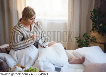 Happy Overweight Woman Reading Book And Having Breakfast In Bed At Home.