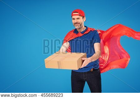 Happy Handsome Delivery Man In Red Superhero Cape And Cap Carrying Cardboard Box. Professional Deliv