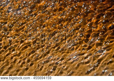 Peat-colored Shallow Water Of River Close Up. Peat River Shallow Clear Water Shimmering At Bright Su