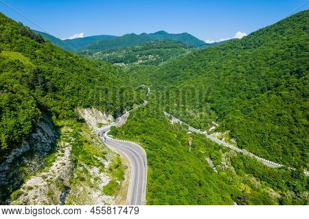 Winding Road Is Meandering Between Blue Sea And Green Mountains. Aerial View Of Car Driving Along Th
