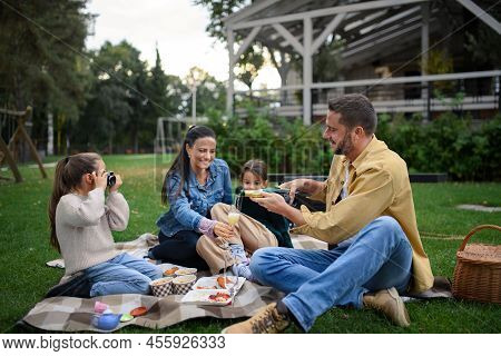Happy Young Family Sitting On Blanket And Having Take Away Picnic Outdoors In Restaurant Area.