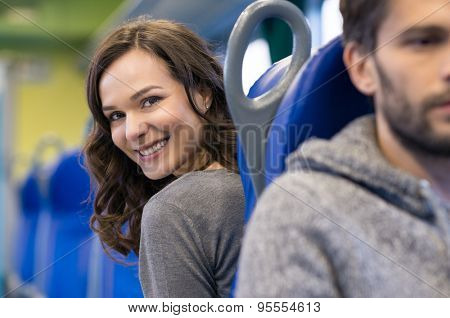 Portrait of a smiling young woman sitting in train. She is a brunette beautiful girl and she is looking back at camera. There are a row of blue seats in the coach.
