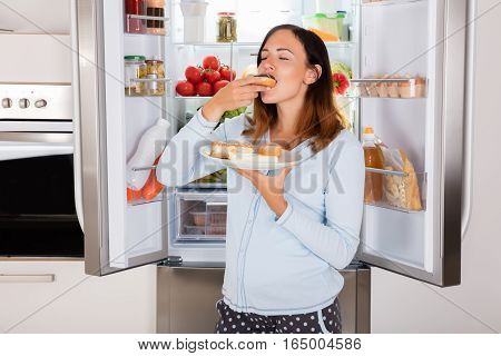 Young Woman Enjoy Eating Donut From Plate Near Refrigerator In Kitchen