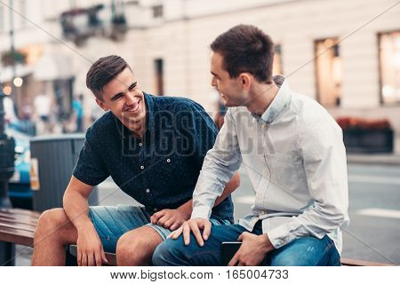 Two smiling young friends enjoying a converseation together while sitting on a bench on a city street