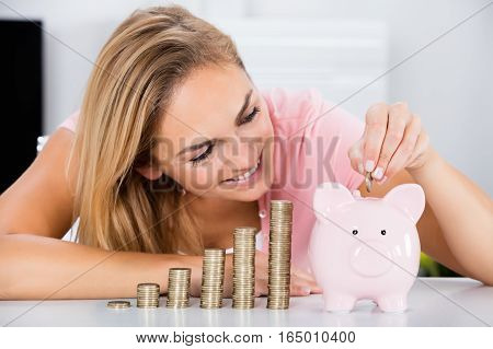 Young Happy Woman Inserting Coin In The Piggybank Near Stack Of Coins On Desk