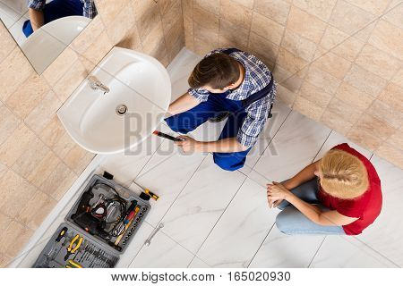 Woman Looking At Young Male Plumber Lying On Floor Fixing Sink In Bathroom