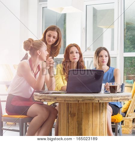 Group of four pretty businesswomen working togeather with new startup project using laptop computer in modern loft. People and teamwork concept - happy creative team in office. Casually dressed female colleagues talking in a meeting room.
