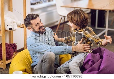 Come to me. Joyful bearded man expressing happiness while caressing his son sitting on hassocks