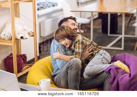 Happy family of two. Joyful young bearded man sitting on hassocks expressing happiness while hugging his son