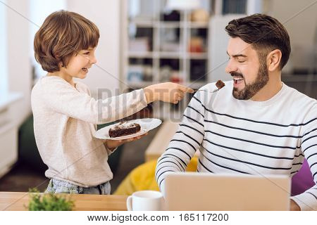 Family look. Young father working with laptop eating piece of cake that gives him his son