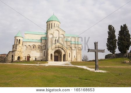 Cathedral of the Dormition or Kutaisi Cathedral more commonly known as Bagrati Cathedral against blue sky background. Bronze cross in foreground.
