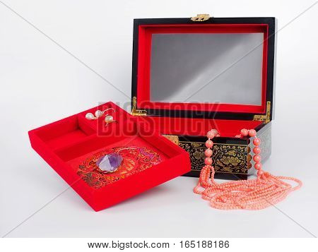 Picture of the opened jewel-box with red fit-out and amethyst pendant and pearl earring . Wooden jewel-box with pink coral bead necklace on white background. Side view.