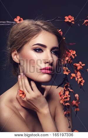 Beautiful young woman with make-up and loose hairdo holding artificial sakura branch with orange flowers. Beauty shot on dark background. Copy space.