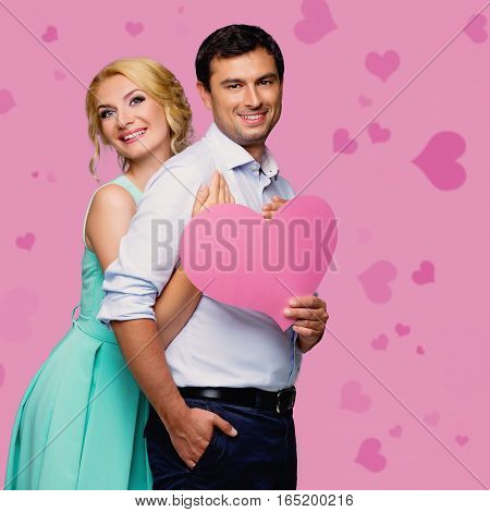 Beautiful young couple holding big pink heart. Blond woman in mint dress and handsome man in shirt and trousers standing on pink background. Valentine's day concept. People in love. Copy space.