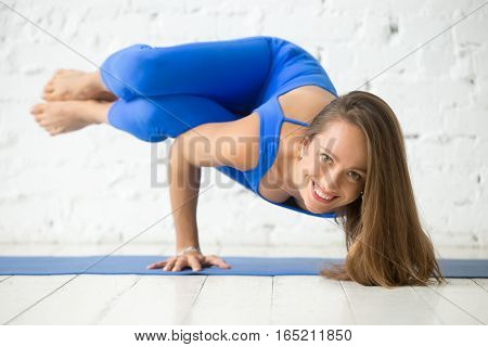 Portrait of young attractive smiling woman practicing yoga, standing in Side Crane exercise, Parsva Bakasana pose, work out wearing sportswear blue suit, full length, white studio, looking at camera