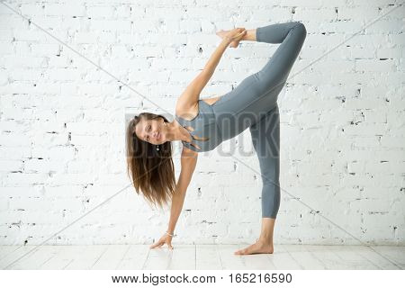 Young attractive smiling woman practicing yoga, standing in Half Moon exercise, Ardha Chandrasana pose, working out, wearing sportswear, grey suit, indoor full length, white loft studio background, looking at the camera