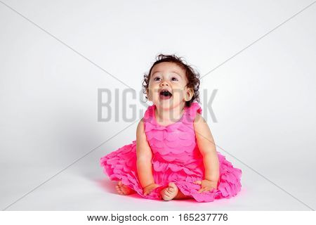 A happy Native American baby girl sits on a white background. She is wearing a pink dress and leans forward with a huge excited open mouth smile.
